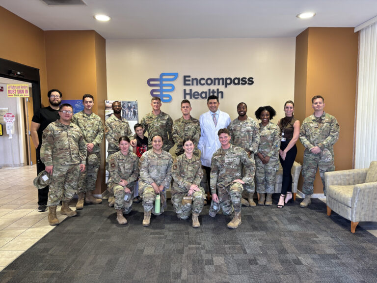 Group of soldiers in camouflage uniforms posing with a few civilians in a lobby in front of an Encompass Health sign and logo.