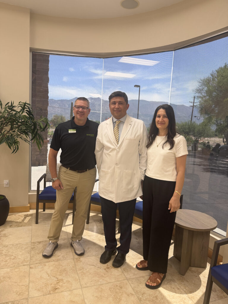 Three adults stand together indoors, smiling for a photo beside a large window with a mountain landscape outside; the center person wears a white coat.