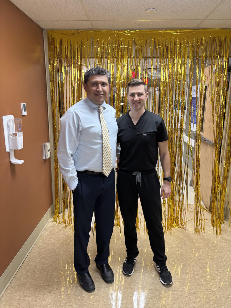 Two men stand in a hallway in front of a gold fringe backdrop, one in a light blue shirt and striped tie and the other in black scrubs, smiling at the camera.